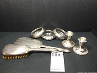 Overall view of sterling vanity mirror, brush, four sterling rimmed glass coasters, and a pair of short silver candle holders arranged on a dark surface.