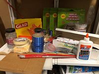 Craft supplies including yarn kits, glue, masking tape, Glad garbage bags, and other assorted items arranged on shelving.