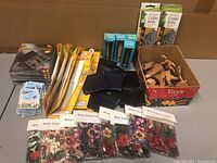 Assorted gift ware laid out on a table including floral wine glass coaster packs, snap coin purses, bookmark magnets, small kitchen tools, thermometer packaging, and a box of wooden shaped cedar pieces.