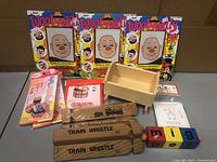 Overall view showing all items included in the lot: Wooly Willy pads, origami kits, wooden cradle, spelling block, wooden train whistles, wire puzzle, and origami paper packs.