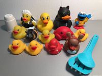Photo showing entire assortment of bathtub toys on a gray surface against white background including various rubber ducks, cookie monster, bee, moose, red octopus, white cup and blue rake toy.