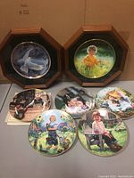 Photo shows seven collector plates, two in wood octagonal frames and five unframed plates. The plates feature children and nature scenes, some with flowers and animals.