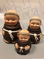 Front view showing three smiling monk figures in brown robes with white rope belts; one small with holes on head, two larger with solid heads.