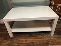 White IKEA LACK coffee table shown from front angle with visible lower shelf and legs on wood floor.