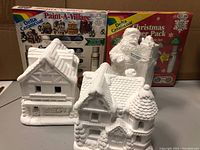 Two unpainted ceramic houses and an unpainted Santa figure with two unopened Delta Ceramcoat paint packs in the background