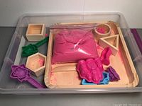 Pink kinetic sand in bag with assorted molds in clear plastic sandbox container.