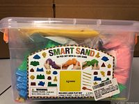 Full view of the clear plastic storage box with Smart Sand label, showing purple and orange sand bags and plastic molds inside.