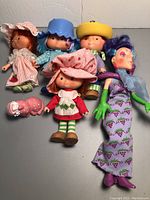 Six dolls arranged on a gray surface including five Strawberry Shortcake dolls and one purple-haired doll in a grape-themed dress along with one small cat figurine. Pink shoes placed separately.