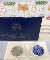 Photo showing 1973 Eisenhower silver dollar coin in plastic sleeve with blue envelope and description card in background, holiday gift bag also visible.