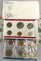 Two sealed plastic mint sets containing six US coins each laid out with a titled envelope showing wear and a holiday gift bag below, showcasing the packaging and contents of the lot.