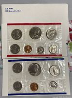 Two sealed plastic mint sets laying on a surface, showing all coins in each set, at least two envelopes with red and blue trim visible