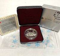 Coin mounted in clear plastic holder inside a hinged black and red velvet display case, with inspection tag, cardboard sleeve, and decorative snowflake plastic bag nearby.
