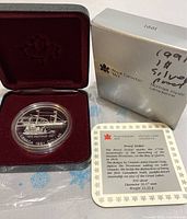 Silver proof dollar coin encased in plastic inside red velvet-lined box, with cardboard box and information card beside it.