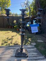 Front view of the three arm metal candelabra showing ornate base and arms holding three candles. Outdoor natural lighting highlights the tarnished metal requiring polishing.