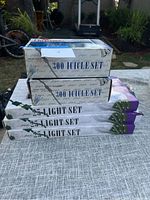 Stacked boxes of Christmas lights including two boxes labeled '300 Icicle Set' and three labeled '25-Light Set' displayed outdoors on fabric surface.