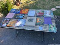 Top-down photo showing all 14 coffee table books laid out on a rustic wooden table outdoors. Various sizes and cover designs visible.