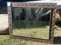 Full front view of rectangular mirror outside leaning upright with visible metal frame and glass surface reflecting outdoor trees and grass.