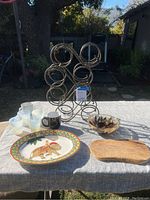 Overview of metal wine rack, drinking glasses, black mug, decorative bowls, plate, and wooden cutting board on table outdoors.