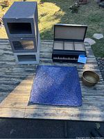 Photo of three shoeboxes stacked, black jewelry box open showing compartments, blue glass tapestry/plaque, and bronze bowl displayed on wooden table.