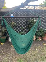 Full view of green single-seat hanging hammock suspended from a tree branch in an outdoor garden with plants in the background.