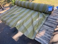 Photo showing the large spool of green and beige striped fabric partially unrolled on wooden surface outdoors.