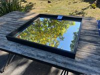 Square framed mirror lying flat on wooden table outdoors, reflecting trees and sky, showing overall condition and size.