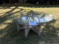 Close-up side angle showing the clear round glass top resting on a wooden base with a circular and cross beam structure outdoors on grass.