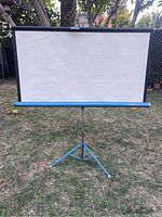 Full front view of vintage Da-Lite movie screen set up outdoors on blue tripod stand, screen fully extended.