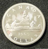 Obverse side of the 1965 Canadian silver dollar coin showing two Indigenous people and a European settler in a canoe with trees in the background and the year 1965.
