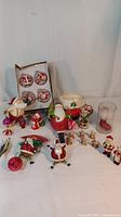 Full lot view including ceramic boot planter, box of glass Christmas ornaments, various Santa figurines, and other Christmas decorations on white cloth background.