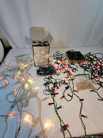 Full view of all Christmas light strings laid out, showing colored bulbs on dark and white cords, vintage push-on flower covers, and new boxed blue lights.