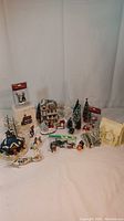 Wide view of Christmas village items including buildings, figurines, trees, and a light cord on a beige background