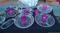 Overall view of glass serving platters, purple candles in clear candle holders, and stemware glasses on black surface.