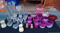Full view of the collection showing various glass items including tall clear vases, crystal stemware, heart-shaped candle holders, two small teacups with gold designs, salt and pepper shakers, frosted purple glasses, red footed bowl, and pink ribbed vase.