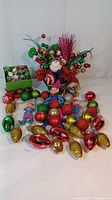 Three colorful knee hugger elves with plastic faces sitting among a large selection of assorted vintage blown glass Christmas ornaments and holiday floral picks.