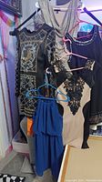 Five women's dresses on hangers shown hanging together including embellished black dress, beige dress with black lace applique, blue halter dress, silver draped neckline dress, and black dress with metallic cuff detail.