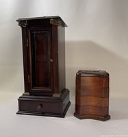 Front view of The Bombay Company tall cabinet-style wooden jewelry box alongside stacked curved multi-tiered jewelry box in dark polished wood and lighter wood finishes respectively.