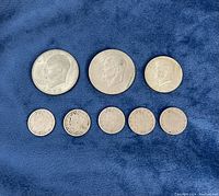 Top view of 8 US coins placed on a blue cloth, showing two Eisenhower dollars, a Kennedy half dollar, and five Liberty Head nickels.
