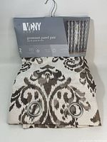 Two grommet curtain panel pairs by VCNY Home with off-white and brown pattern. Panels are folded and displayed with packaging showing brand and product info.