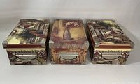 Front angled view of three decorative boxes with vintage-style artwork on lids and metal handles on front sides. Metal corner protectors are visible.