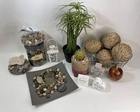 Boho themed home decor items arranged including faux plant, twine balls on metal stand, jars and dish of pebbles with candle holder, small wooden quote blocks, wooden apple decor