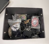 Top view of the black box containing several plastic sealed bags with various cords including black, beige, red, and multicolor cords alongside leather strips and necklace templates.