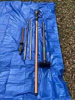 Multiple lawn equipment items laid out on a blue tarp outdoors on grass and leaves. Including poles, stamper, limb trimmer head, and a handheld trimming tool.