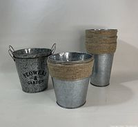 Front angle photo of 7 galvanized buckets showing 3 styles: 1 bucket with handles and print, 3 medium buckets with burlap and rope, 4 tall buckets with rope wrap