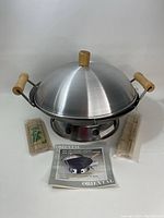 Metal wok and steamer with dome lid and wood handles, two packs of wooden skewers, and original instruction manual displayed in a white backdrop.