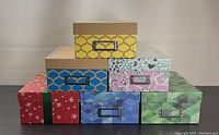 Six decorative rectangular photo storage boxes stacked in two rows, showing various patterns including red snowflake, blue geometric, yellow trellis, pink floral, green leafy, and blue diamond pattern. Metal label holders visible on the front of each box.