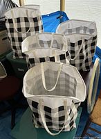 Four large matching black and white plaid laundry bags with handles shown standing upright on plastic containers and a chair, bags open at the top