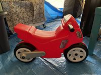 Side angle view revealing the full red plastic body, black handlebars, and white wheels. Motorcycle shape for toddlers.
