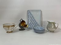 Seven vintage and antique kitchen items displayed side by side on a white surface including cups, plates, a platter, a pitcher, and a duck-shaped bottle opener.