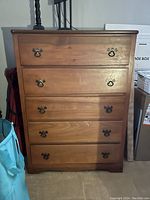Front view of the wooden dresser showing all five drawers and metal handles. The dresser is placed against a wall with some items around it.